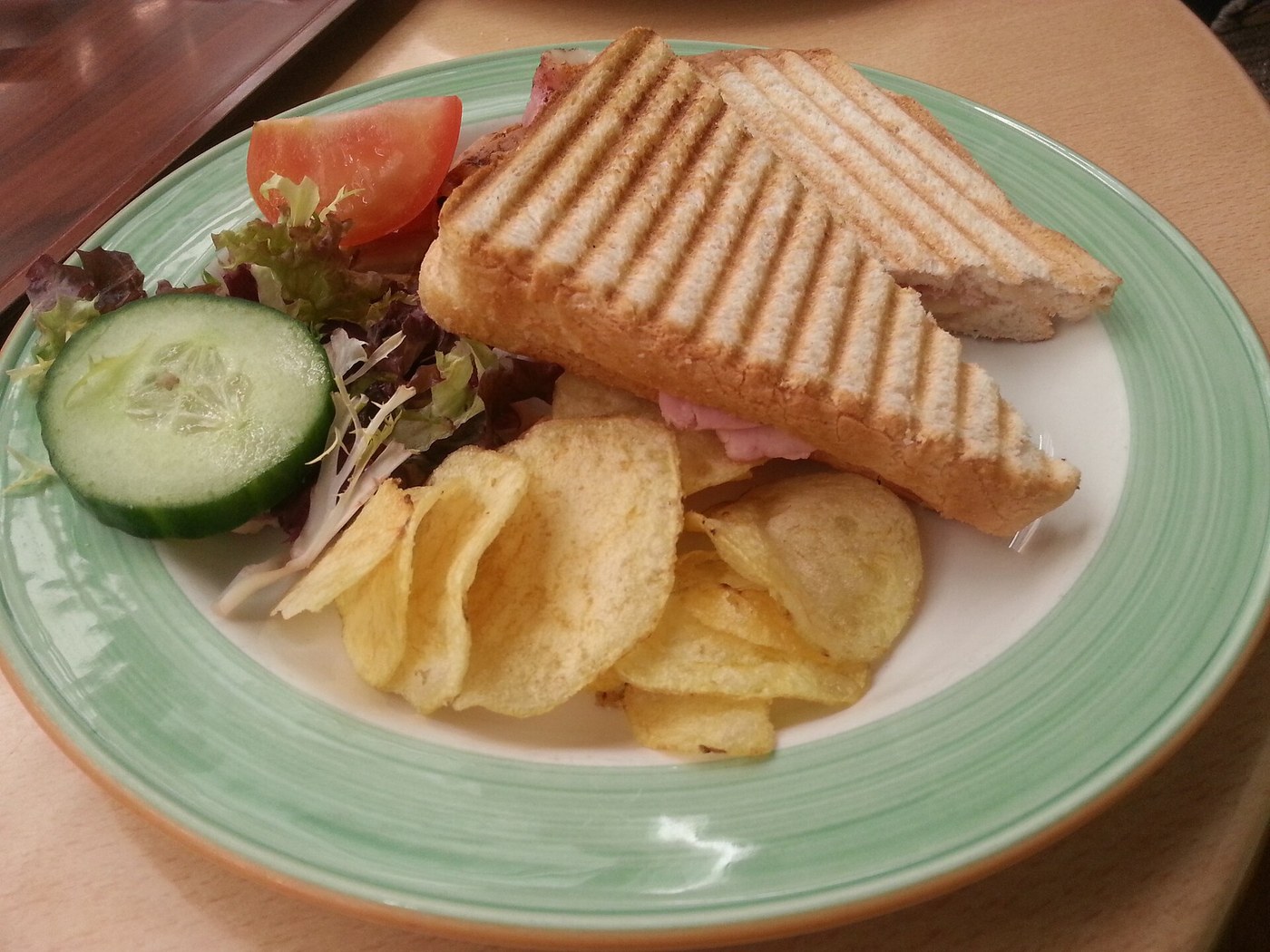 Grillezett sonkás-sajtos melegszendvics félbevágva, chips, uborka, paradicsom és saláta körettel, zöld szegélyű tányéron